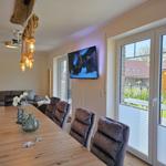 Esszimmer mit Holztisch, Sitzgelegenheiten und TV an der Wand. Blick auf den Garten durch Glastüren.