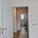 Flur mit Holzboden führt in das Badezimmer. Weiße Türen und Wände.