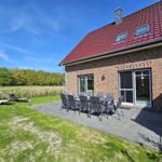 Haus mit Terrasse, Garten und Außentisch mit Stühlen. Hintergrund: Wald und blauer Himmel.