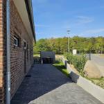 Pflasterweg neben Backsteinhaus mit Garten und Wald im Hintergrund.