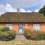 Rotes Backsteingebäude mit Strohdach, blauer Tür und Blumenbeete.
