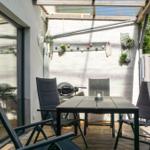 Terrasse mit Tisch, Stühlen und Grill unter Dach. Hintergrund: Holzzaun und Pflanzen.