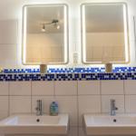 Zwei Waschbecken mit beleuchteten Spiegeln und blau-weißem Fliesenspiegel.