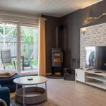 Wohnzimmer mit Sofa, Fernseher und Holzofen. Blick auf Garten durch Glastür.