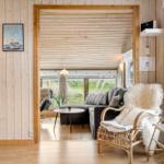 Wohnraum mit Holzwänden, Sofa und Sessel. Fenster zeigen Grünfläche. Wandbild zeigt Boot.
