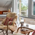 Sitzgelegenheit mit Felldecke und Kissen vor großem Fenster. Im Hintergrund Schlafbereich mit orangefarbenen Kissen.