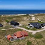 Villa mit rotem Dach und schwarzen Häusern am Strand mit Dünen und Windrädern im Hintergrund.