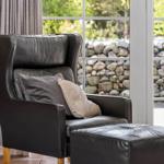 Schwarzer Ledersessel mit Kissen und Fußhocker vor großem Fenster mit Steinmauerblick.