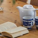 Holztisch mit Teekanne, Tassen und offenem Buch.