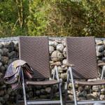Zwei Stühle mit Decke stehen vor Steinmauer und Bäumen.