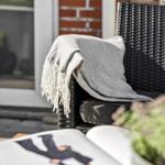 Schwarzer Rattanstuhl mit grauer Decke und offenes Buch auf Tisch.