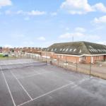 Tennisplatz mit Umzäunung und angrenzenden Gebäuden unter blauem Himmel.