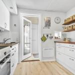 Küche mit weißen Schränken, Holzoberflächen und integriertem Ofen. An der Wand hängen Körbe und ein Wanduhr.