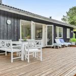 Terrasse mit weißem Tisch und Stühlen, Sitzbank und Pflanzen vor einem Haus mit dunkler Fassade.