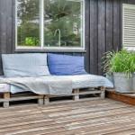 Außenliegende Matratze auf Paletten mit Kissen und Decke vor dunklem Holzhaus.