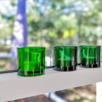 Drei grüne Teelichter stehen auf einem weißen Fensterbrett vor einer verschwommenen grünen Landschaft.