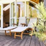 Zwei Liegestühle auf Holzterrasse vor Glasfront mit Blick ins Haus.