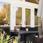 Terrasse mit Tisch, Stühlen und Sonnenschirm. Hintergrund: Glasfront und Pflanzen.