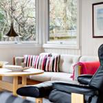 Ein Wohnzimmer mit Sofa, Tisch und schwarzer Ledercouch. Durch Fenster blickt man auf Bäume.