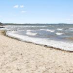 Strand mit Sand und Wellen, Küstenlinie mit Felsen und Bäumen im Hintergrund.