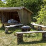 Holzschuppen mit Matratze, Grill und Bänken aus Baumstämmen im Garten.