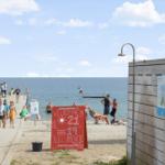 Strand mit Badesteg, Menschen und rotem Willkommensschild.