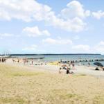 Strand mit Steg, Sonnenbadenden und Segelyachten im Hintergrund.