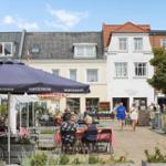 Außenbereich mit Tischen, Sonnenschirmen und Passanten vor historischen Gebäuden.