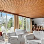 Wohn- und Essbereich mit weißen Sesseln, Holzdecke und großen Fenstern zum Garten.