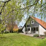 Haus mit Terrasse, Garten und rotem Dach unter Bäumen.