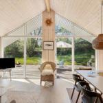 Wohnraum mit Holzwand, großem Fenster und Essbereich. Draußen Terrasse mit Sonnenschirm.