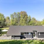 Ein dunkel verputztes Haus mit Holzveranda und Garten. Umgeben von Bäumen und einem weißen Sonnenschirm.