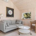 Wohnzimmer mit Sofa, Rattan-Stuhl und Glas-Tisch. Holzwände und Fenster mit Blick auf Grün.