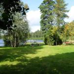 Grüner Rasenbereich mit Bäumen und Blick auf See. Picknicktisch und Stein im Vordergrund.