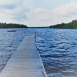 Holzsteg führt über das Wasser zu einem Boot. Wald umgibt den See.