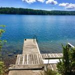 Holzsteg führt zu einem See mit klarer blauer Wasserfläche und grüner Waldlandschaft im Hintergrund.