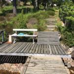 Holzbrücke mit Bank über Bach, umgeben von Grün und Steinen.