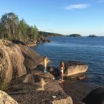 Zwei Kinder stehen auf Felsen am Wasser. Im Hintergrund sind Bäume und Inseln zu sehen.