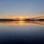 Sonnenuntergang über einem ruhigen See mit Spiegelung im Wasser und Wald am Horizont.