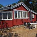 Rotes Holzhaus mit Terrasse und Esstisch. Umgeben von Bäumen unter blauem Himmel.