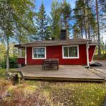 Rotes Holzhaus mit Terrasse am See, umgeben von Bäumen und einem kleineren Anbau.