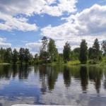 See mit Spiegelung, Bäume und roter Hütte am Ufer unter blauem Himmel mit Wolken.