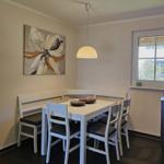 Essbereich mit weißem Tisch, Stühlen und Wandbild. Fenster und Küchenbereich sichtbar.