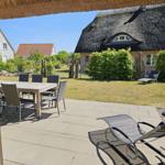 Terrasse mit Tisch, Stühlen und Liegestuhl. Blick auf Garten und strohgedecktes Haus.