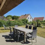 Terrasse mit Esstisch und Stühlen unter strohgedecktem Dach. Grünfläche und Häuser im Hintergrund.