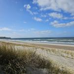 Strand mit Dünen und Wellen unter blauem Himmel mit weißen Wolken.