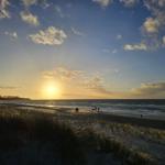 Sonnenuntergang am Strand mit Wellen und Menschen in der Ferne.