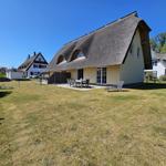 Haus mit Strohdach, Garten, Terrasse und Gartenmöbel unter blauem Himmel.
