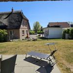 Terrasse mit Liegestuhl und Tisch. Hintergrund: Haus mit Strohdach und Garten.