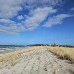 Strand mit Dünen, See und Dorf im Hintergrund unter blauem Himmel mit weißen Wolken.